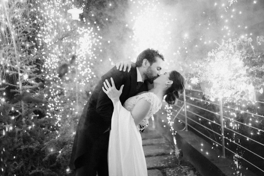 foto en blanco y negro de los novios con la luz e las bengalas frías de fondo y dándose un beso apasionado