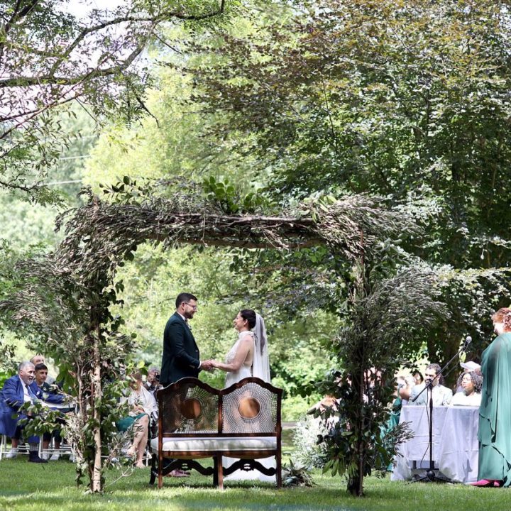 foto de boda la finca de A Quinta da Auga en donde se ve un arco de plantas con troncos, a los novios y el banco de ceremonias habitual de A Quinta da Auga
